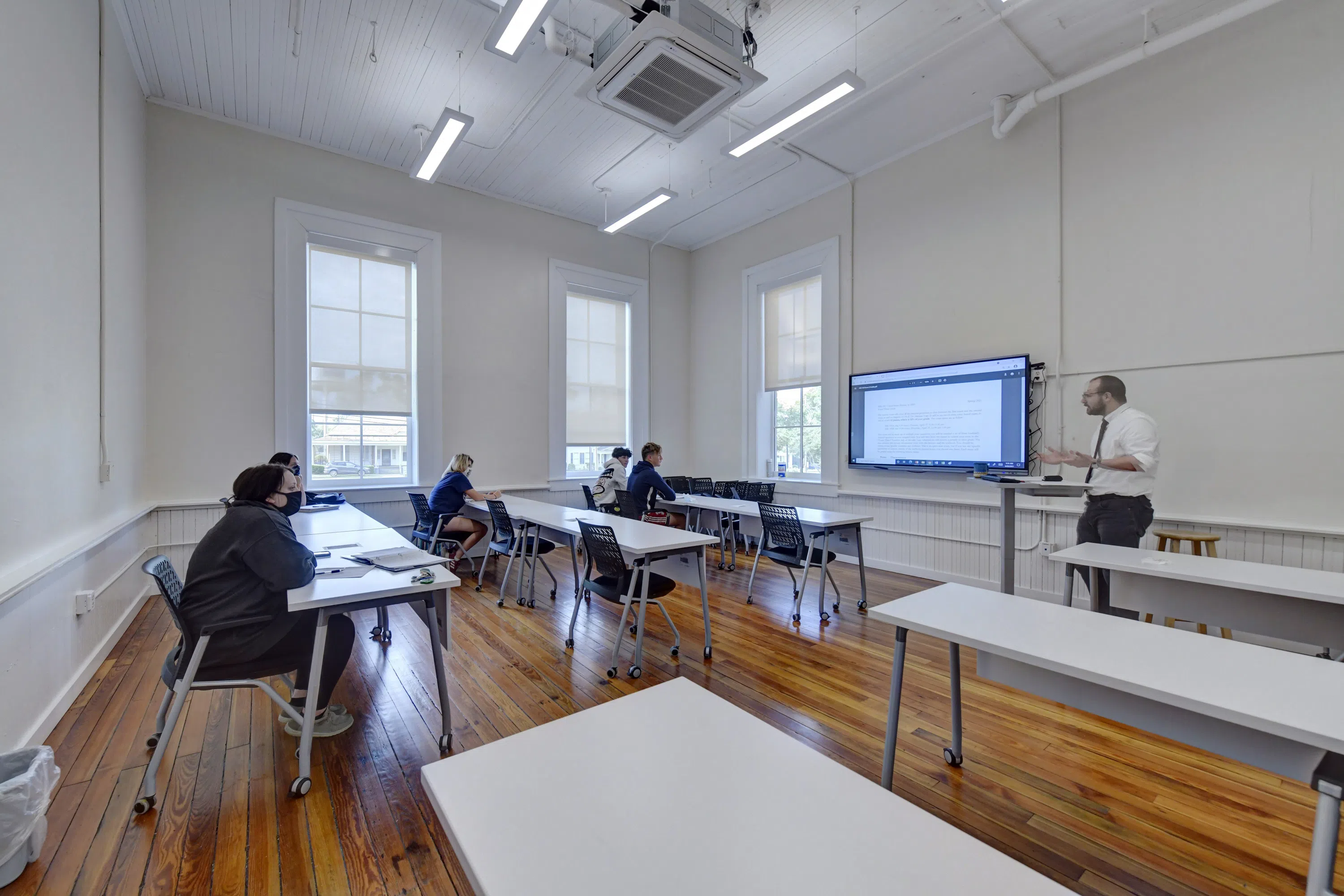 Photo of students in classroom in Staples Hall seated at desks looking at professor with large screen monitor on wall. 