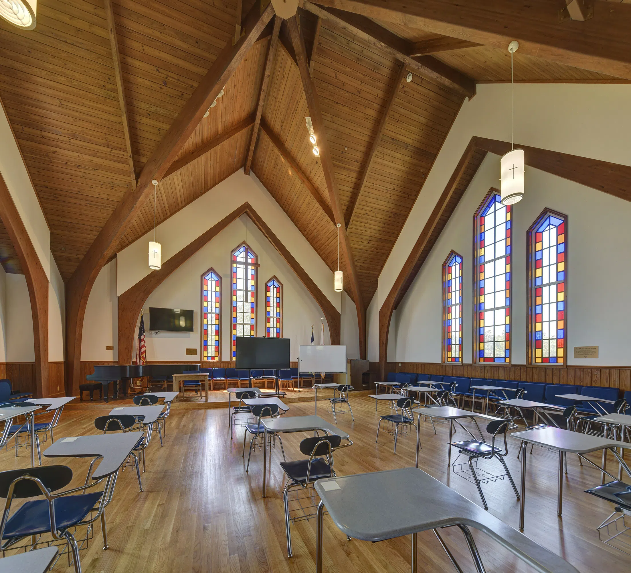 Desks setup inside Jones Chapel sanctuary with stained glass windows , mounted TV, large whiteboard, American flag, piano, and organ. 