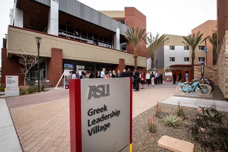 Gray and Maroon sign says ASU Greek Leadership Village in front of a courtyard filled with people next to a two-story building with a sign that says ASU Greek Leadership Village Community Center. 