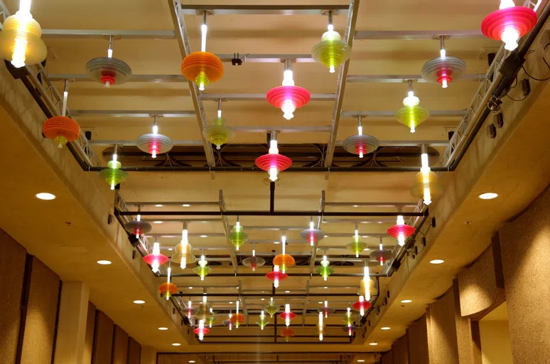 View of a ceiling in a walkway that has a series of lights with pink, green, orange and yellow discs around them. 