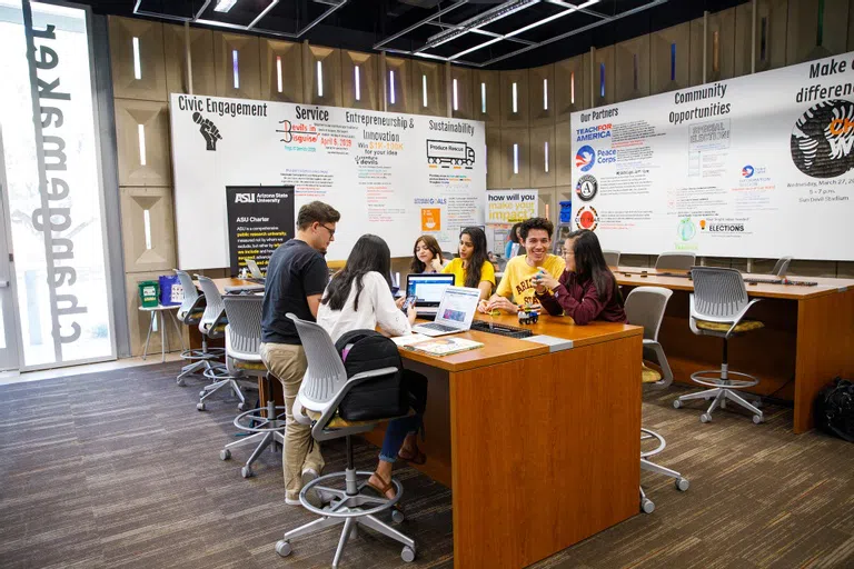 Six students sit around a high top table with a couple laptops open. Around them are white boards covered with writing about upcoming events and initiatives. 