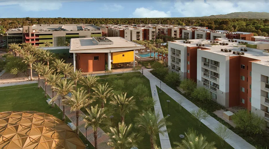 a palm tree-lined walkway cuts through a lawn to a series of four-story apartment buildings that are white with brick-red accent walls. In the center is a brick-red and yellow two story building and a community pool. 