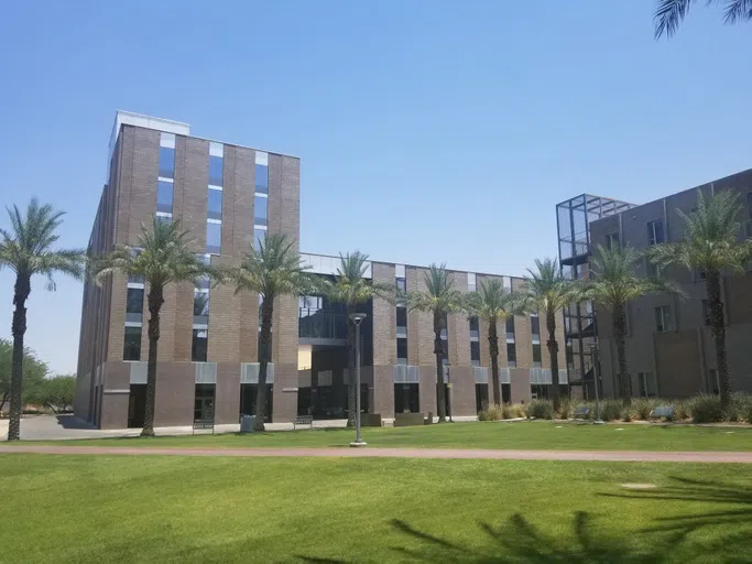 A lawn and line of palm trees are in front of seven-story and four-story cinderblock buildings connected by a second-floor bridge. 