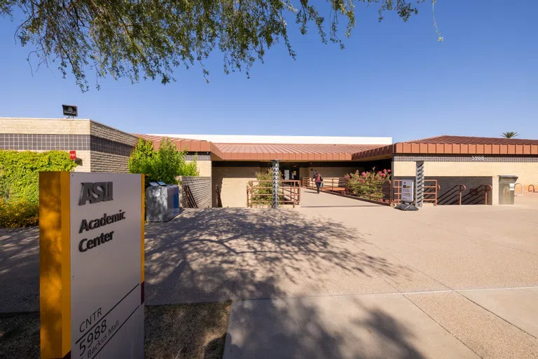 Metal sign reading "Academic Center" stands in front of a brown-tone building where a woman is walking up the center pathway lined by metal railings.