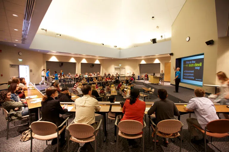 A presenter stand in the middle of the room with a floor to ceiling screen behind him saying The Sun Devil Way. The room is full of people sitting at tables in concentric half circles around the presenter.