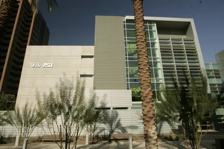 Trunk of a palm tree in front of concrete and glass building with ASU and UA logos on it