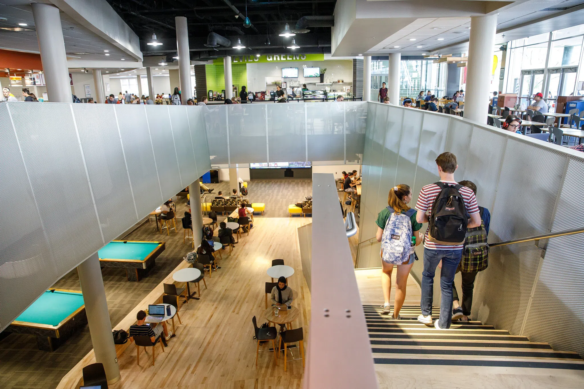 Three people walk down a stairway from a first floor dining area into a lower level recreation area with seating and billiard tables.