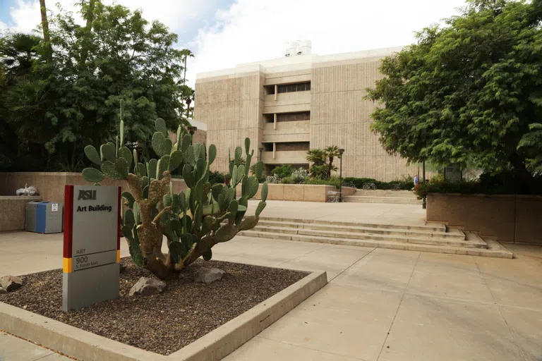 A sign saying Art Building sits next to a large prickly pear cactus in a walkway in front of stairs leading up to a four-story concrete building.