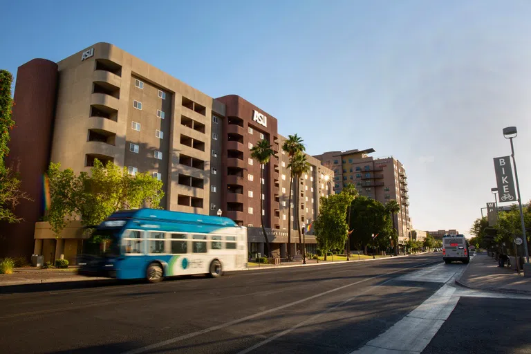 A blue and white bus passes by a seven-story stuccoed building. The center of the building is a mauve color and either side is beige. At the top of the building is a sign that says ASU.