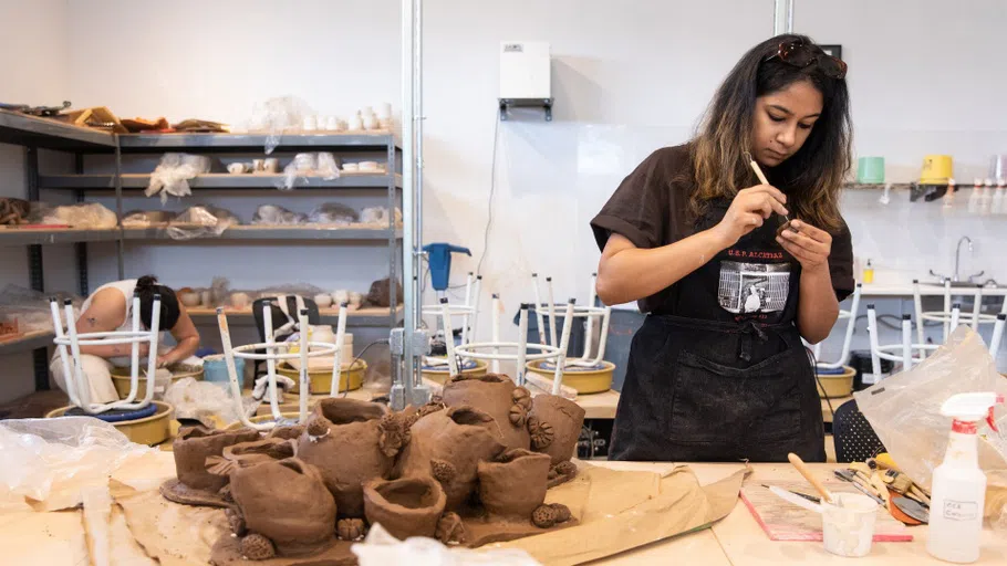 A student standing up works on her sculpture, while a student in the background works on the wheel. 