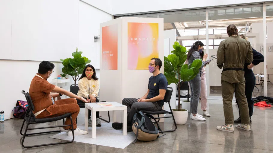 Four students sit and discuss something around a small white table with plants around them; others stand and converse off to the right.