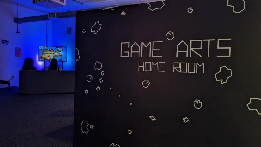 A dark, carpeted space is illuminated by purple and blue light; a mural on the front wall reads "Game Arts Homeroom" and is decorated with puzzle pieces and a mouse icon.