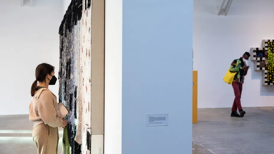 A student looks closely at a large textile mounted to a wall; the space behind shows a large gallery space with another viewer taking in other works.