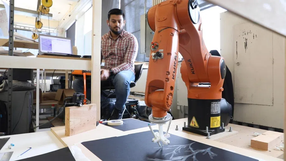 An orange robotic arm draws a tree-like form on black paper.