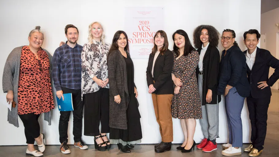 A group of presenting VCS graduates stand with CCA professors near vinyl that reads 2019 VCS Spring Symposium.