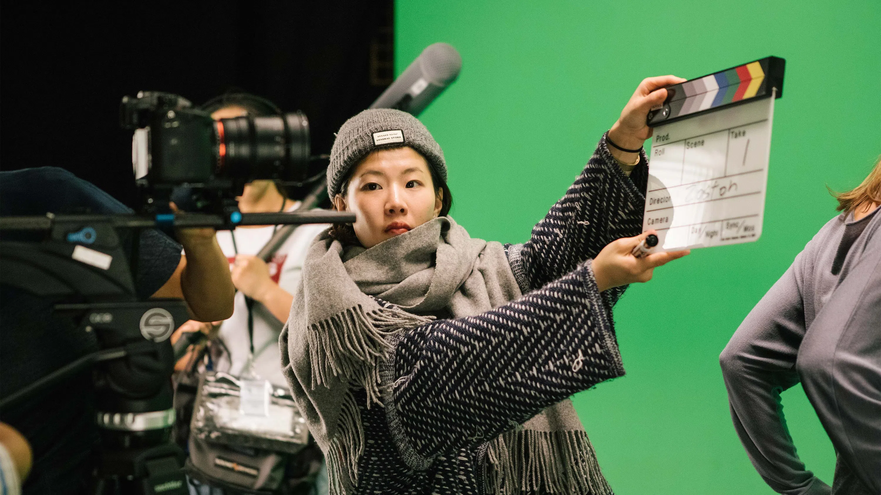 A student claps a slate in front of a DSLR camera, signifying the first take of a scene in a green-screen environment.