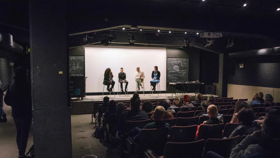 A wide shot of four people in conversation on stage in a dark lecture hall filled with attendees.