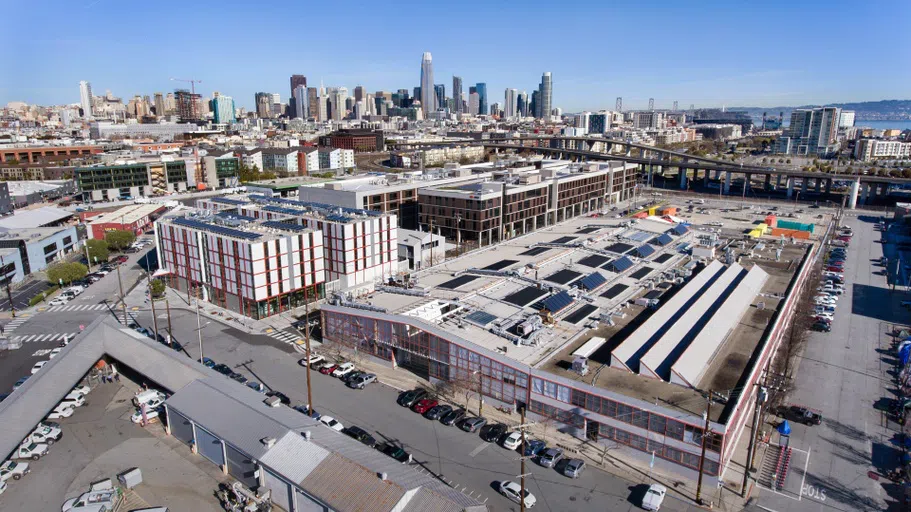 An aerial view of CCA's backlot with colorful chalk drawings of abstract shapes, painted containers, potted plants, and picnic tables decorating the concrete area. The main building recedes in the background.