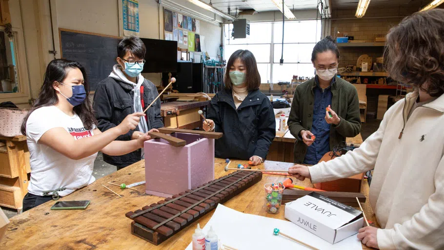 Five students play with hand-made marimas around a wooden table in the Bench Room.