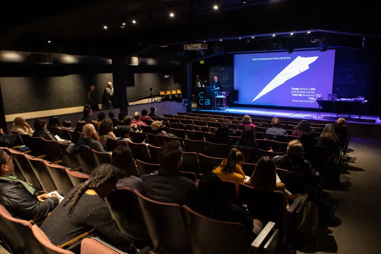 A dark space with a purple slide up on a screen; someone stands at a podium with CCA vinyl.