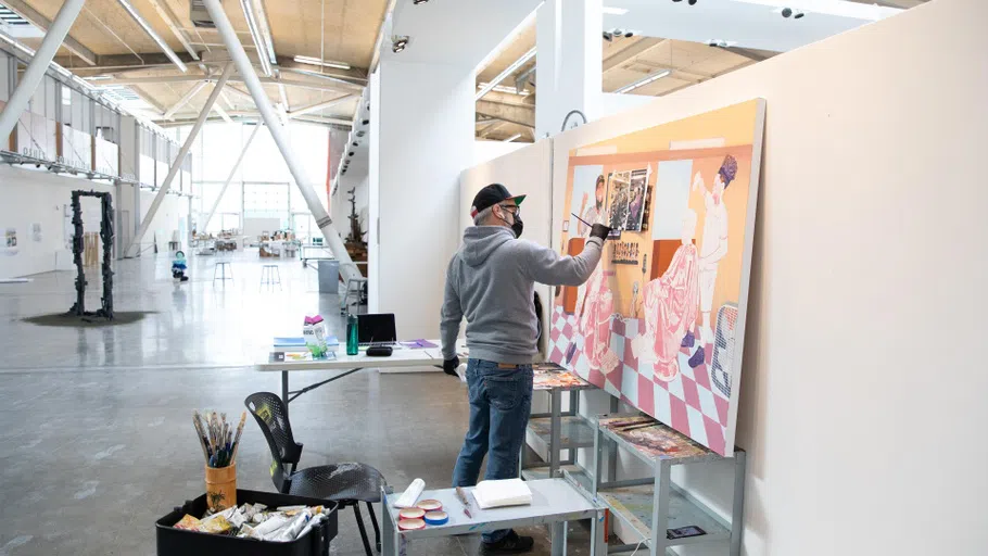 A student holds a paintbrush to a larger canvas painting. 