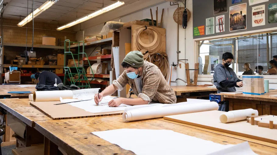 A student sketches on large pieces of paper on a wooden table. In the background, another student tapes and measures a project.