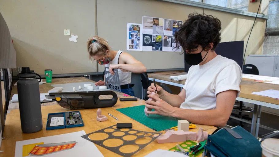 Two seated, masked students prototype individual projects at long desks in the Industrial Design studio.