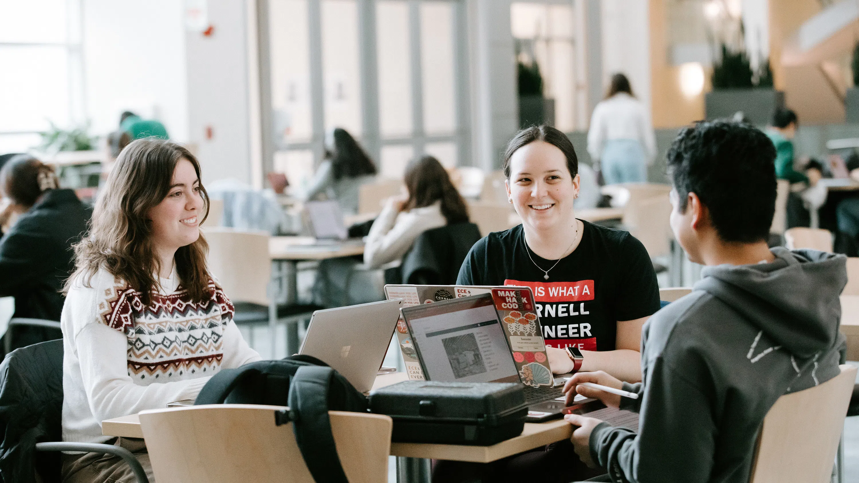 Three Cornell Engineering students hang out at a table in the atrium of Duffield Hall, with large glass windows in the background. 