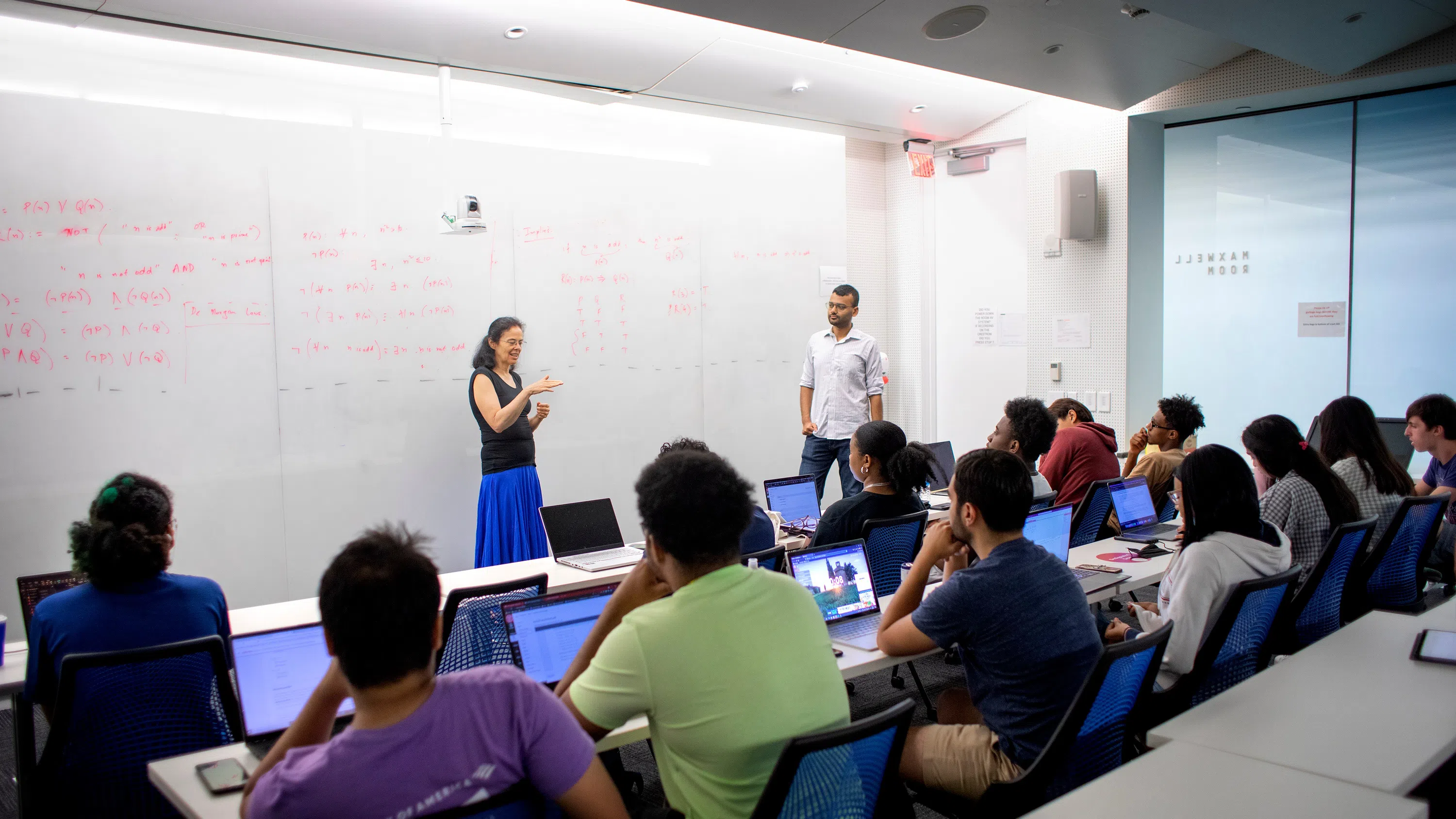 A professor, standing at left center, leads a class while two rows of students look on.