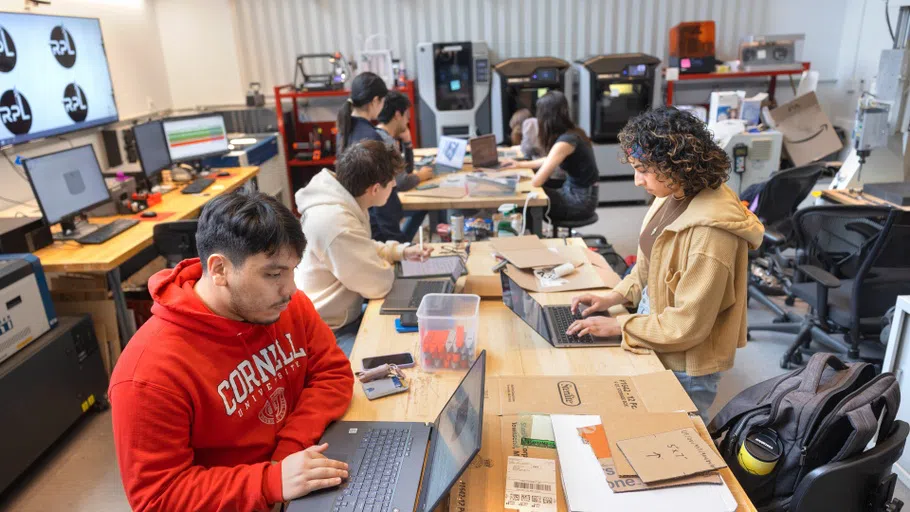 Six students sit at a long table. In front of them are laptops and other tech, as well as pieces of cardboard and other tools. In the background are several high-end 3-D printers.