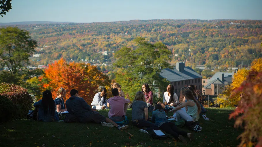 About a dozen students sit and lie in circle while studying. In the background are trees in full autumn foliage.