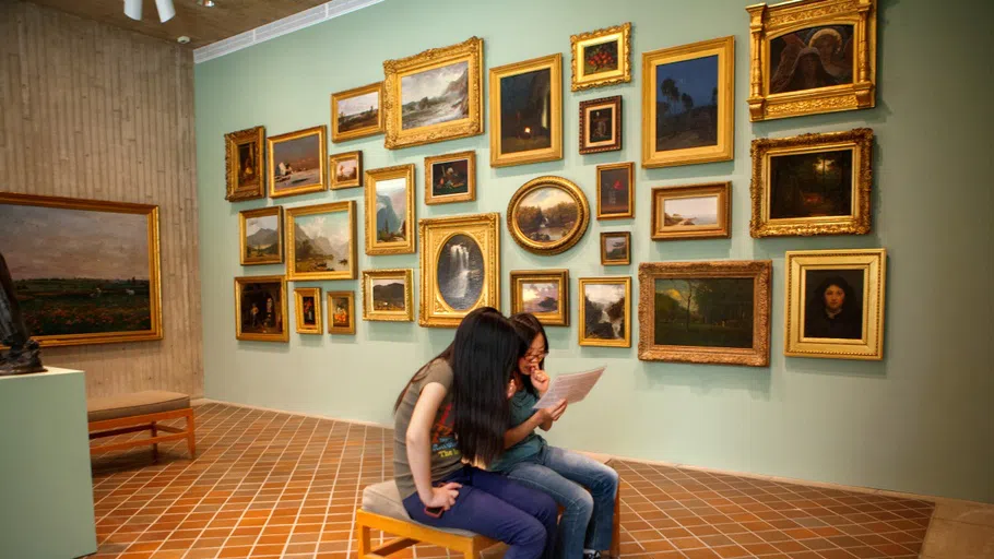 Two visitors to the Johnson Museum, sitting on a bench in the middle of a gallery, read more about the exhibit. Behind them are about two dozen small paintings mounted on a wall.