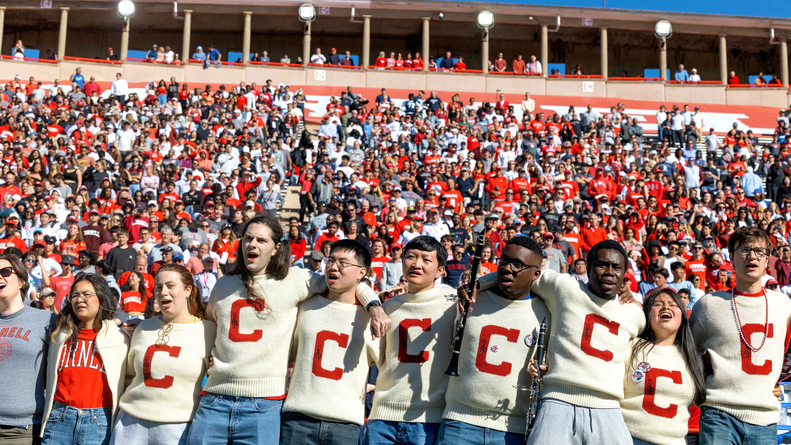 Eight Cornell students are locked arm in arm. Each is wearing a white sweater with a red letter C on the front. Behind them are stadium stands filled with people. 
