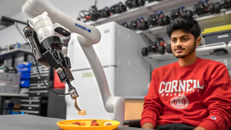 A student, right, wearing a red Cornell sweatshirt, watches a robot arm that is holding a fork with a piece of banana.