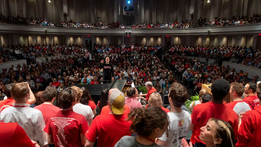 Dozens of performers on stage, seen from behind, perform in front of an audience of hundreds in Bailey Hall. 