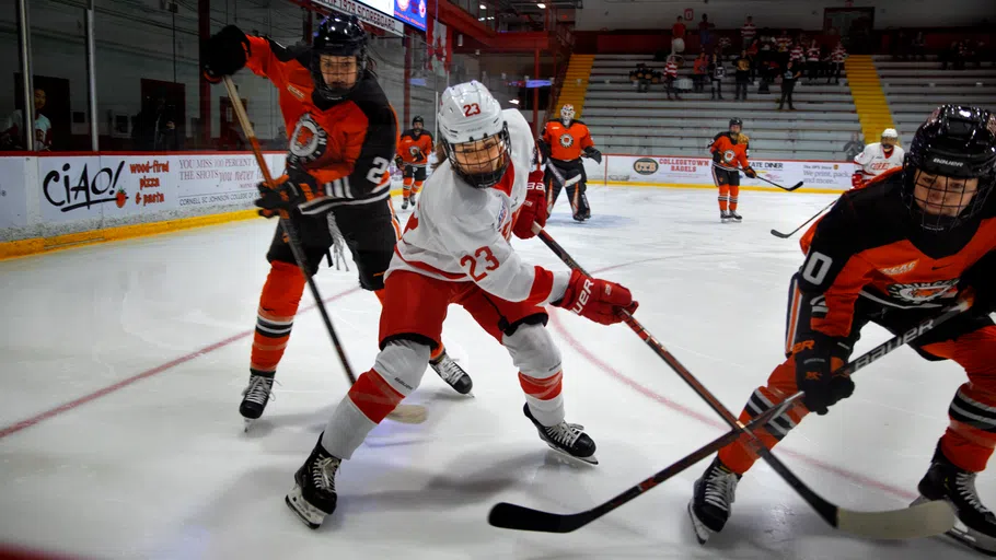 A Cornell women’s hockey player in white, center, is flanked by two opponents dressed in red and black colors.