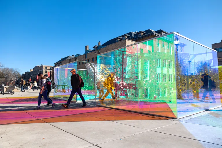 Two individuals walking near the PolyForm, whose exterior colored-glass walls reach about seven feet high and reflect onto nearby pavement.