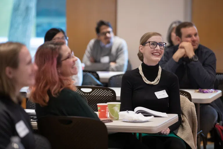 Two women at a round ballroom table listen to a speaker off-screen.