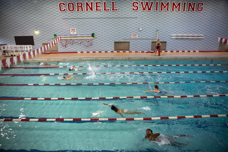 Students take a swim test in an Olympic-sized pool. Each lane is separated by red-and-white lane markers, and the words “Cornell Swimming” is seen on the far wall. 