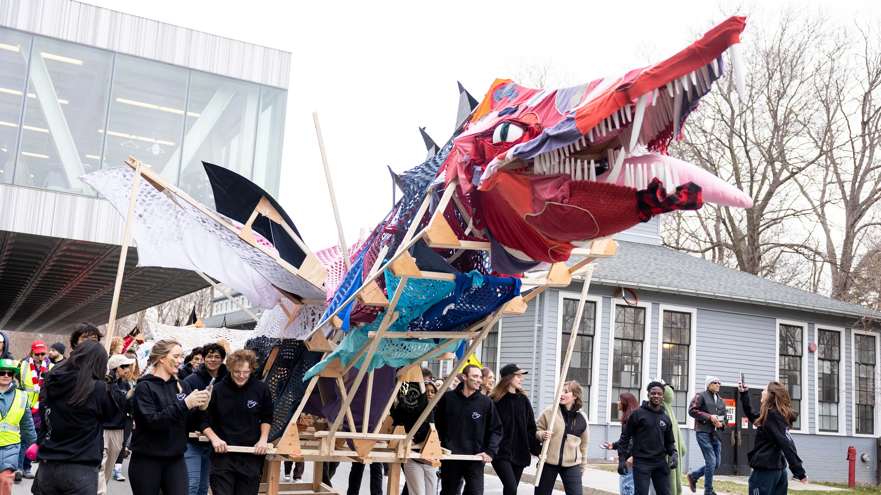 Several dozen students parade a large dragon art project, created from wood, fabric and other materials. It has a red head and very large white teeth.