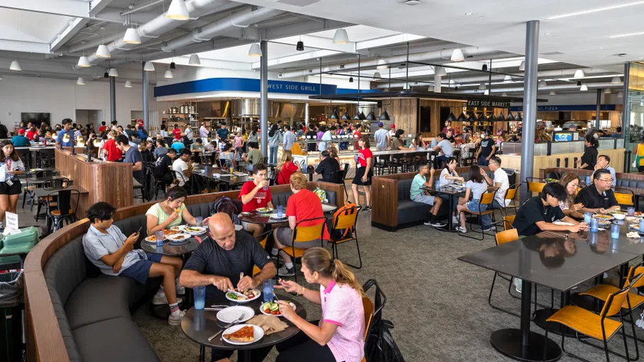 A wide shot of a dining hall, where dozens of students and families eat together. In the background are signs that read “West Side Market” and “East Side Market.”