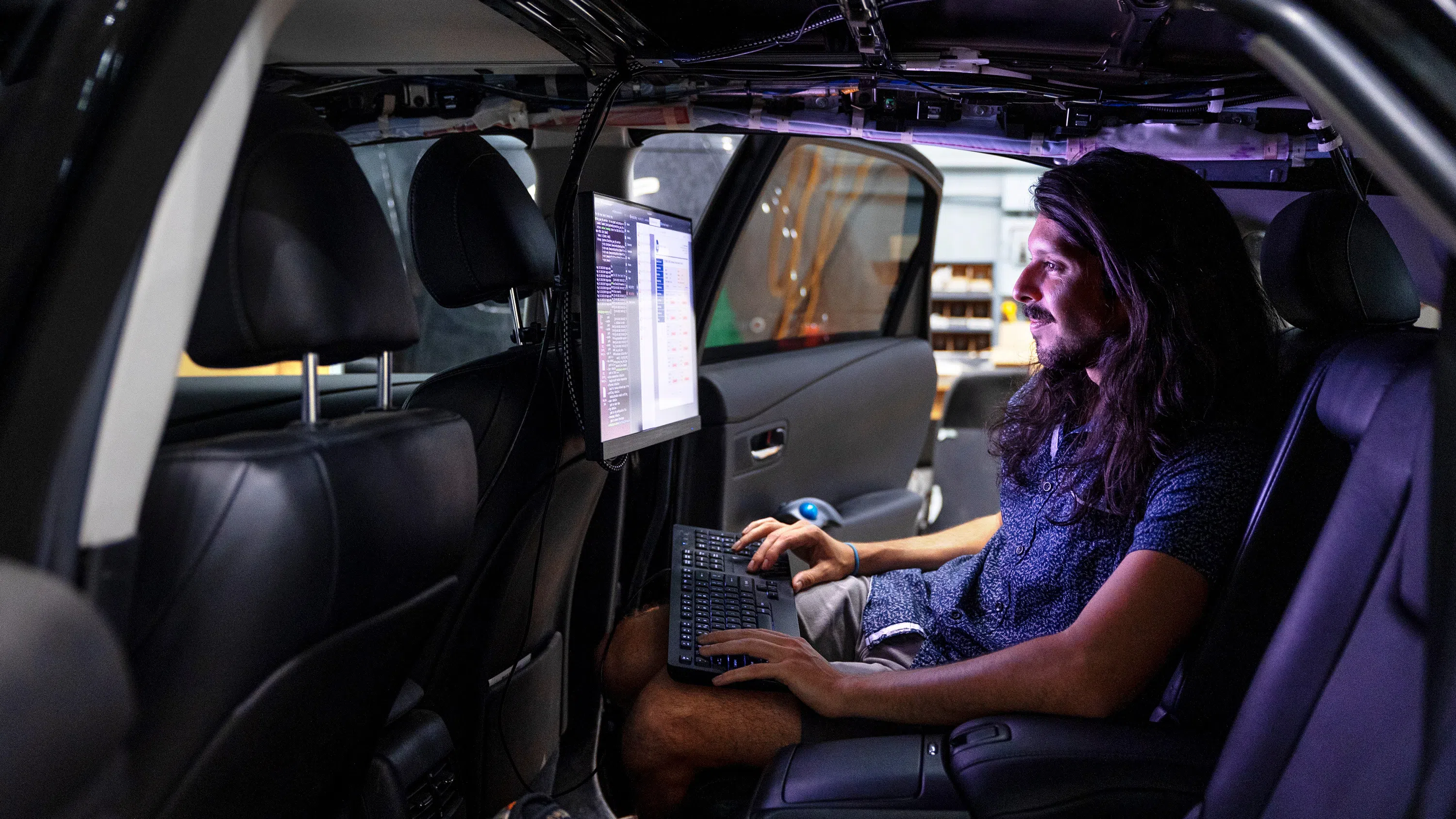 A man with a beard and long hair sits in the back seat of a car. In front of him is a bright screen and a keyboard on which he is typing.