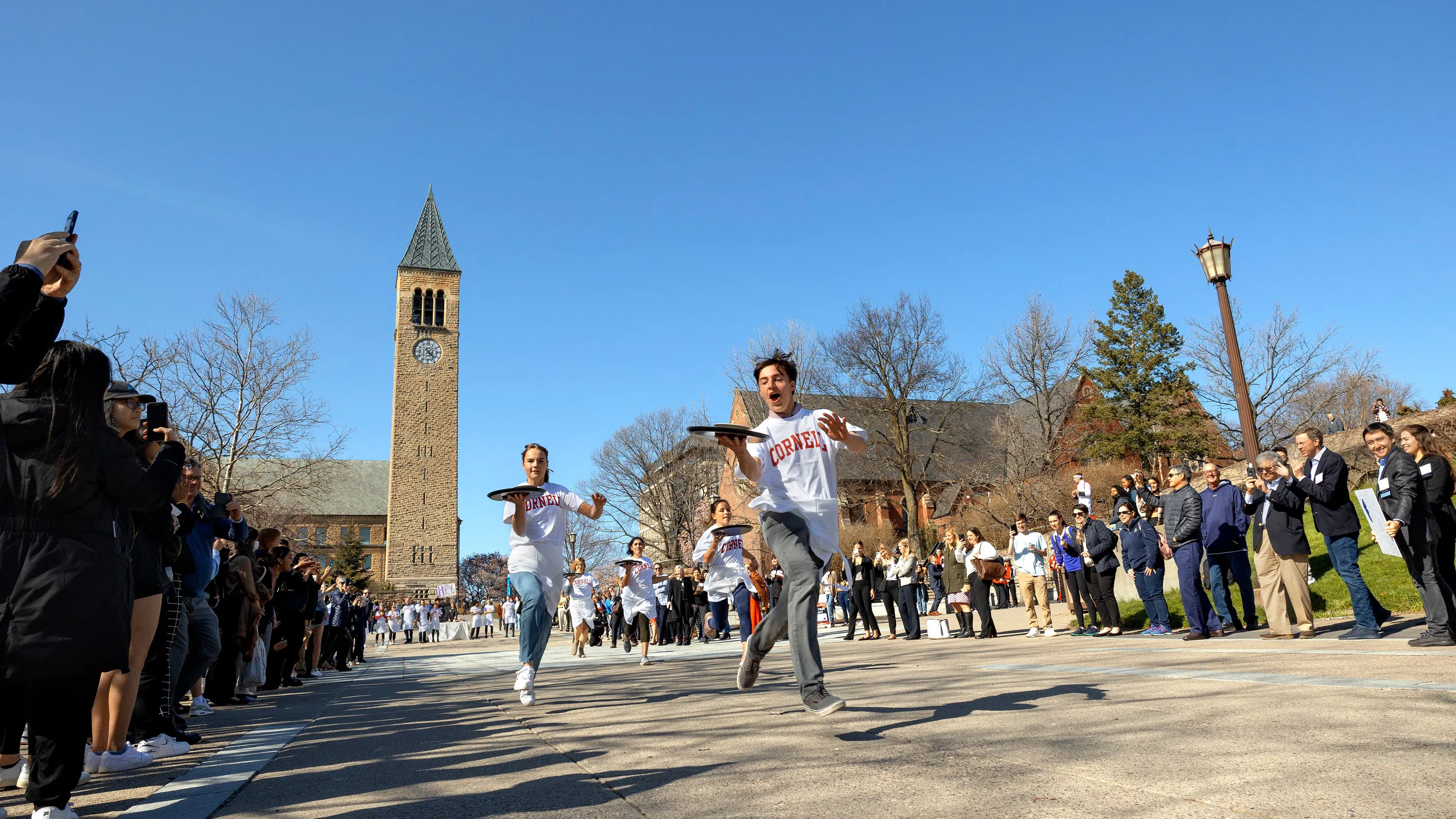 Five students wearing white aprons race across a plaza while holding server trays of soup. Onlookers cheer on and take photos. McGraw Tower is in the background against a blue sky.