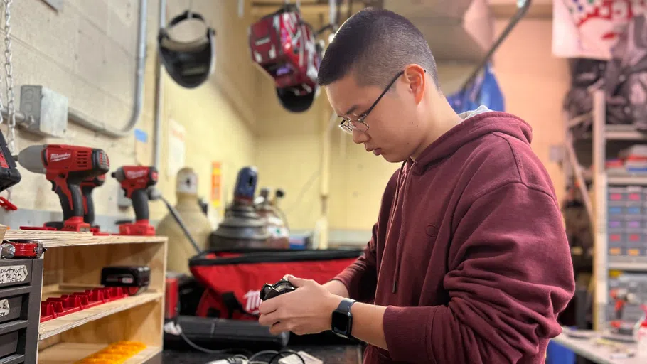 A male student wearing a maroon hoodie sweatshirt works on a project at a workbench.