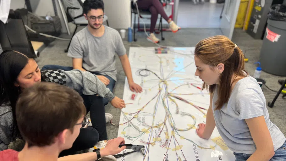 A half-dozen students look at a large poster on the floor, printed with various terms related to the parts of a race car. Many of the terms are connected together by pieces of string.