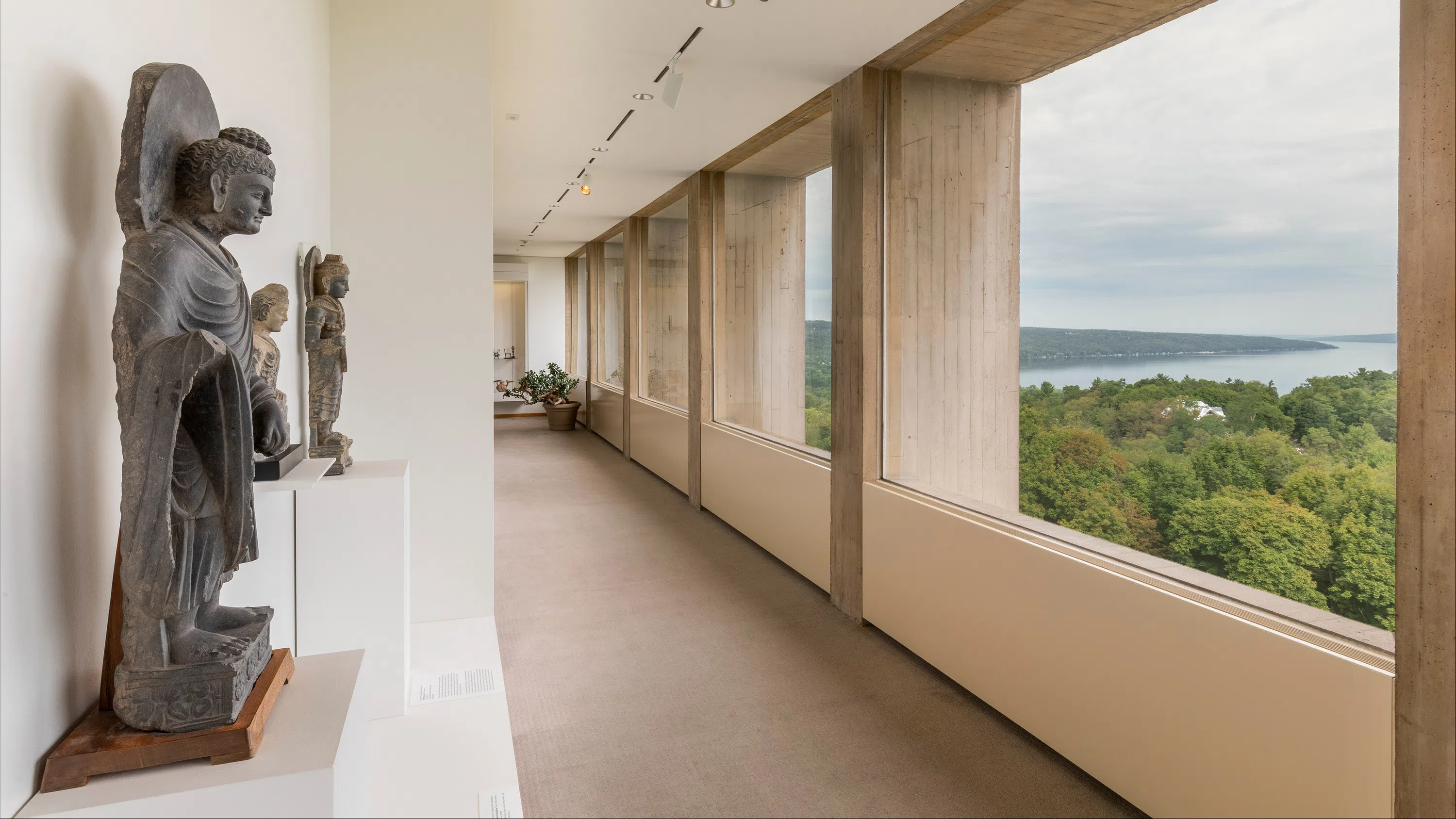 Statues line the left side of a museum hallway; to the right are large glass-pane windows with a wide view of Cayuga Lake.