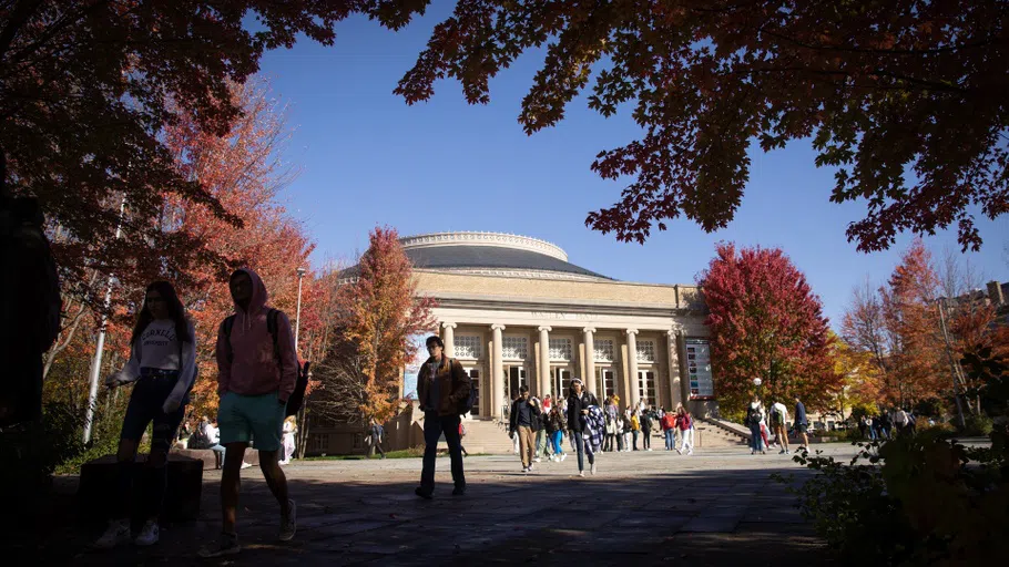 Dozens of students walk across a plaza, with Bailey Hall behind them. The photo also has several trees displaying fall foliage in reds, browns, and yellows.