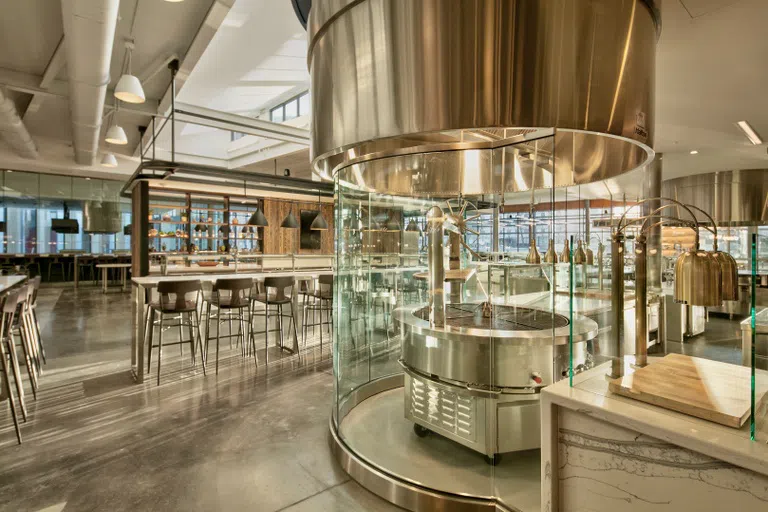 One of the four main serving areas at Morrison Dining. Photo shows sleek, glass-and-steel warming stations, long communal tables with high-top chairs, and windows.