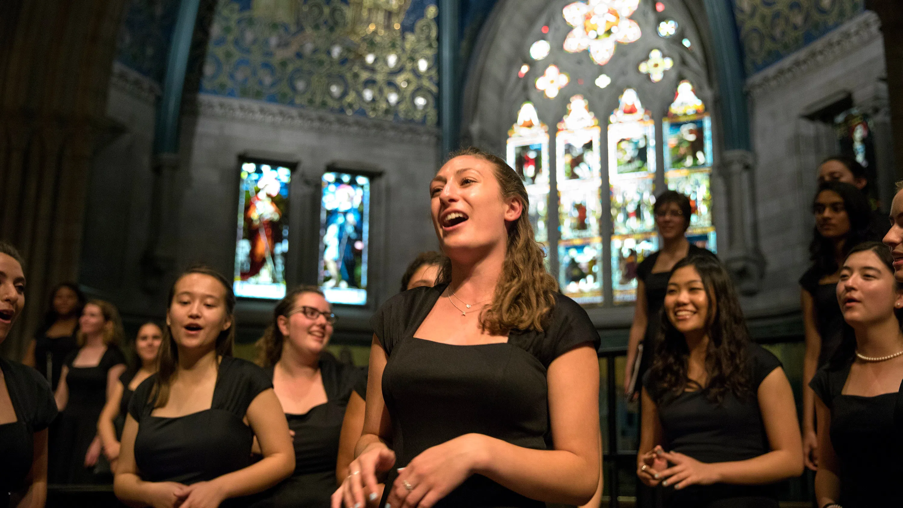 Choir singers perform in Sage Chapel, with multiple stained-glass windows behind them.