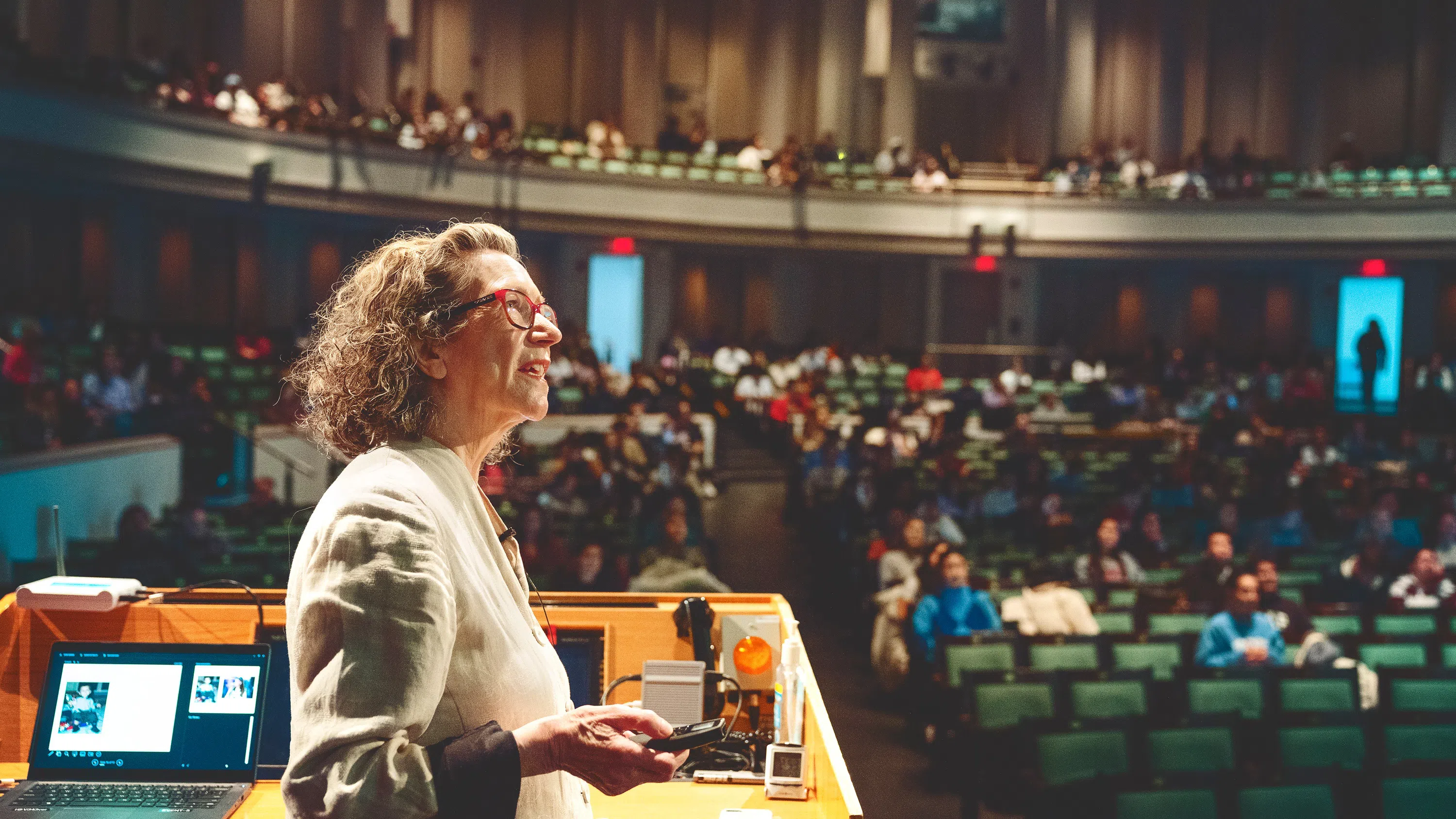 A faculty member, seen at left, looks toward the right as she teaches a class. In the background are dozens of students attending the lecture, including several on a balcony.